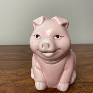 Peut inclure: Une tirelire en céramique rose avec un visage souriant et les yeux fermés. La tirelire est posée sur une surface en bois.