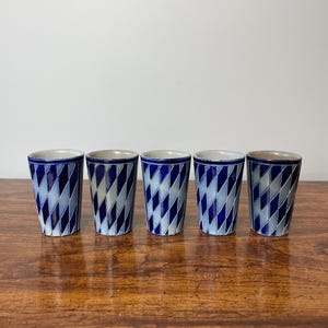 May include: Five ceramic shot glasses with a blue and white geometric pattern. The glasses have a cylindrical shape and are decorated with a repeating diagonal design. They are displayed on a wooden surface.