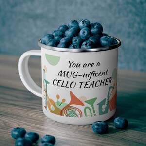 May include: White enamel mug with a colourful musical instrument design and the text "You are a MUG-nificent CELLO TEACHER". The mug is filled with blueberries.