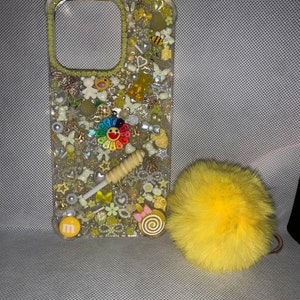 May include: A clear phone case with a yellow, white, and rainbow design. The case is covered in small, colorful embellishments, including stars, hearts, and flowers. A yellow pom-pom keychain is attached to the case.