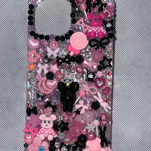 Può includere: Una custodia per telefono rosa e nera con una varietà di ciondoli, tra cui orsi, fiocchi, stelle e cuori. La custodia è decorata con glitter e strass.