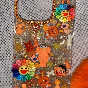Op de afbeelding: Een transparante telefoonhoesje versierd met kleurrijke harscharms, waaronder bloemen, beren en sterren. De hoes is versierd met een rand van oranje strass.