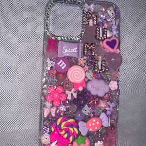 Op de afbeelding: Een transparante telefoonhoesje versierd met roze, paarse en witte versieringen. Het hoesje heeft een verscheidenheid aan bedels, waaronder snoep, bloemen, harten en een Hello Kitty-personage. De tekst "Sweet" en "Mumu" is zichtbaar op het hoesje.