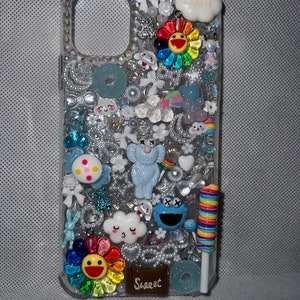 Op de afbeelding: Een transparante telefoonhoesje versierd met kleurrijke strasssteentjes, glitter en verschillende bedels, waaronder een regenboogbloem, een blauw monster, een wolk, een lolly en een kleine olifant. Het hoesje heeft ook het woord "Sweet" erop.