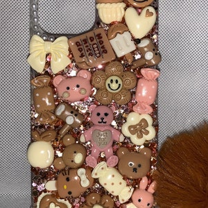 Op de afbeelding: Transparante telefoonhoes versierd met diverse bruine en roze bedels, waaronder teddyberen, bloemen, strikken en snoepjes. De hoes heeft een glitterachtergrond en de tekst "Have a nice day".