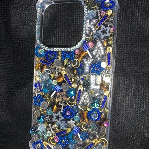 Op de afbeelding: Transparante telefoonhoes versierd met een assortiment blauwe, zilveren en gouden bedels. De bedels omvatten veiligheidsspelden, kralen, sterren en lolly's. De hoes heeft een transparante rand met iriserende strass-steentjes.