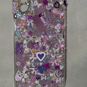 May include: A clear phone case decorated with a variety of purple, white, and silver embellishments, including hearts, flowers, stars, and bows. The case also features a small, purple cloud with the word "Regal" written on it.