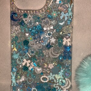 May include: A clear phone case decorated with blue, white, and silver glitter, rhinestones, and charms. The charms include a Hello Kitty character, a blue bear, a blue butterfly, and a blue bow.