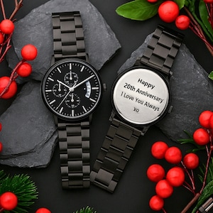 May include: Two black metal wristwatches with link bands. One watch features a black dial with three sub-dials and white markers. The other watch shows a silver-toned back casing engraved with the text: Happy 20th Anniversary I Love You Always XO.