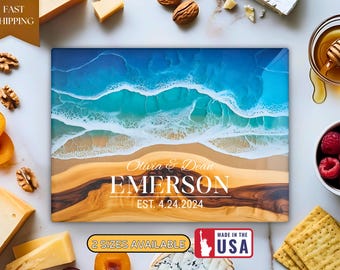 May include: A rectangular cutting board with a beach-themed design. The top features a blue ocean with white waves, a sandy beach, and a wooden base. The text "Olivia & Dean EMERSON EST. 4.24.2024" is displayed in white.
