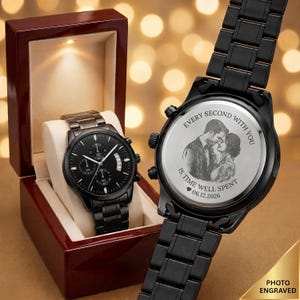 May include: A black metal wristwatch with a link band and a black dial featuring sub-dials. The back of the watch case is engraved with a portrait of a couple, the text "EVERY SECOND WITH YOU IS TIME WELL SPENT," and the date "06.12.2026."