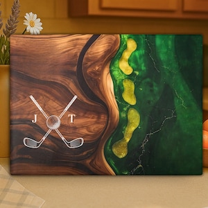 Puede incluir: Una obra de arte de madera con tem&aacute;tica de golf con un dise&ntilde;o &uacute;nico. La obra presenta una combinaci&oacute;n de madera oscura y resina epoxi verde, con un dise&ntilde;o de campo de golf. El dise&ntilde;o incluye palos de golf cruzados, una pelota de golf y las letras "J" y "T".
