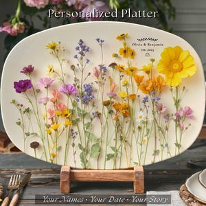 May include: An oval, cream-colored platter with a floral design featuring colorful wildflowers. The platter is displayed on a wooden stand. Text at the top reads "Personalized Platter" and includes names and a date.