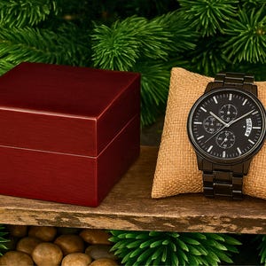 May include: A black chronograph wristwatch with a metal link band displayed on a tan burlap pillow next to a dark red wooden box. The watch face features white hour markers, three sub-dials, and a date window positioned at the three o'clock mark.