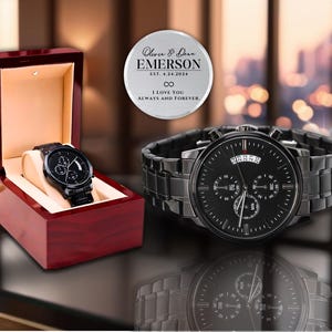 May include: A black chronograph watch with a date window and a black leather strap. The watch is in a red wooden box. A silver engraved disc with the text "Oliver & Deer EMERSON EST. 4.24.2024 I LOVE YOU ALWAYS AND FOREVER." is in the background.