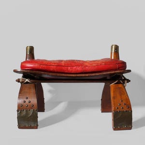 Può includere: Sella di cammello vintage con cuscino in pelle rossa. La struttura in legno presenta dettagli in ottone e borchie metalliche. La sella è progettata con una seduta curva e gambe robuste, mostrando una miscela di materiali e artigianato.