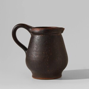 Può includere: Una brocca in ceramica marrone scuro con un manico curvo. La brocca ha un corpo arrotondato e un bordo leggermente svasato. La superficie ha un aspetto strutturato con piccole macchie scure. Il manico è spesso e si curva dolcemente.