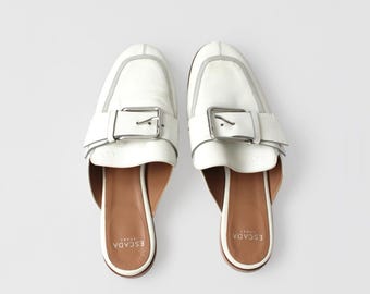 May include: A pair of white leather loafers with a silver buckle and a brown leather insole. The loafers have the text "EScada" printed on the insole.