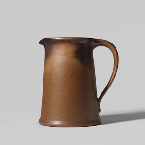 Op de afbeelding: Een keramische kan met een taps toelopend cilindrisch lichaam en een gebogen handvat. Het oppervlak heeft een matte afwerking met een verloop van aardse bruintinten, van lichtbruin aan de basis tot donkerbruin bij de bovenrand.