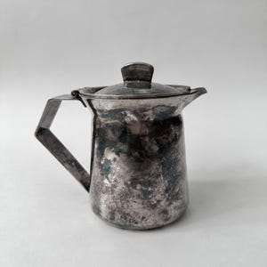 May include: A tarnished silver-toned metal pitcher with a hinged lid and a flat, angular handle. The body of the pitcher has a cylindrical shape with a small pouring spout and a rounded base, showing visible signs of oxidation and surface wear.