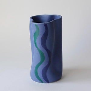 May include: A blue ceramic vase with a wavy design in shades of blue and green. The vase has a unique, curved shape and a matte finish. The vase is a decorative item.