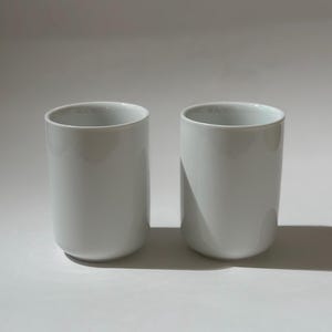 May include: Two white ceramic cups with a cylindrical shape. The cups have a smooth, glossy finish and a slightly rounded base. The cups are displayed on a white surface.