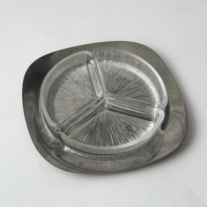 Op de afbeelding: Een vintage serveerschaal met een heldere glazen inzet met drie secties, geplaatst in een geborsteld metalen dienblad. De glazen inzet heeft een radiaal patroon en de schaal heeft een afgeronde vierkante vorm.