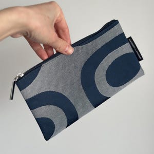 Pode incluir: Uma bolsa retangular com um padrão abstrato azul marinho e cinza. A bolsa tem um fecho de correr e uma pequena etiqueta preta com texto branco. O design apresenta formas curvas em forma de onda.