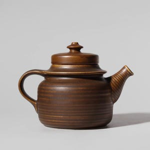 Könnte beinhalten: Eine braune Keramik-Teekanne mit Deckel und gebogenem Griff. Die Teekanne hat einen Ausguss mit gerilltem Design und einen runden Körper mit horizontalen Rillen. Der Deckel hat einen kleinen Knopf oben. Die Teekanne ist warmbraun.