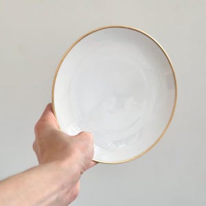 Op de afbeelding: Een witte keramische plaat met een gouden rand wordt tegen een lichtgrijze achtergrond gehouden. De plaat is rond en heeft een eenvoudig, elegant ontwerp. De gouden rand voegt een vleugje verfijning toe aan de verder effen witte plaat.