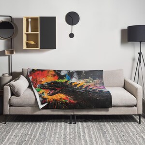 May include: A black and white throw blanket with a colorful abstract design featuring a Godzilla-like monster. The blanket is draped over a gray couch with a throw pillow.