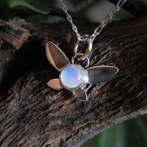 May include: A silver pendant necklace featuring a flower-shaped design with four petals and a central, iridescent opal stone. The pendant hangs from a delicate silver chain. The necklace is displayed against a textured, wooden background.
