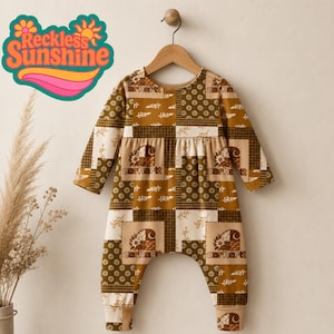 May include: A long-sleeve romper featuring a patchwork design in shades of brown, tan, and cream. The pattern includes floral motifs, checkered squares, and the Carhartt logo. The garment is displayed on a wooden hanger against a neutral background.