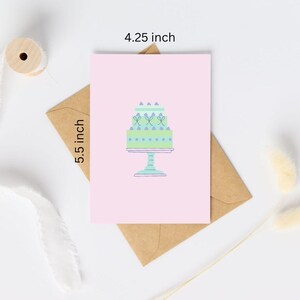 Op de afbeelding: Een roze kaart met een groene en blauwe taart met witte frosting en paarse sprinkles op een witte voet. De kaart is 4,25 inch bij 5,5 inch.