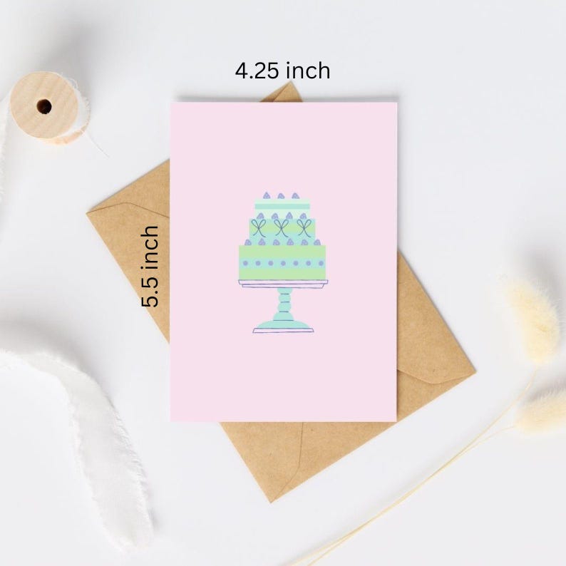 Op de afbeelding: Een roze kaart met een groene en blauwe taart met witte frosting en paarse sprinkles op een witte voet. De kaart is 4,25 inch bij 5,5 inch.