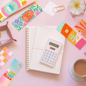 Puede incluir: Una vista cenital de una superficie rosa con varios marcadores de colores, un cuaderno, una calculadora, una taza de caf&eacute; y un calendario. Los marcadores presentan diferentes dise&ntilde;os y citas inspiradoras, como "Trabaja duro, s&eacute; amable", "Eres hermosa, por dentro y por fuera", "Vive tu vida" y "S&eacute; amable con tu mente".