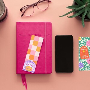 Puede incluir: Una vista plana de un diario rosa con un marcap&aacute;ginas a cuadros, un tel&eacute;fono inteligente, una taza de caf&eacute; y tres marcap&aacute;ginas coloridos con citas inspiradoras. Los marcap&aacute;ginas presentan dise&ntilde;os florales, un patr&oacute;n a cuadros y un personaje de dibujos animados.