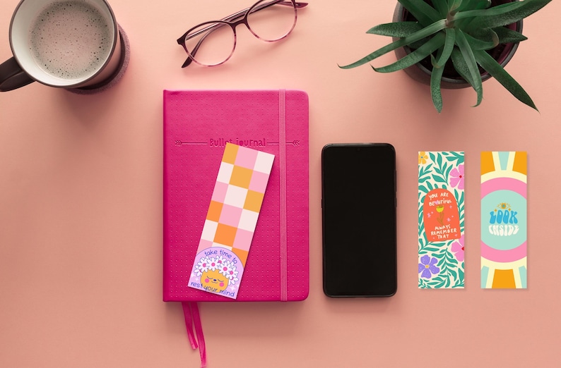 Puede incluir: Una vista plana de un diario rosa con un marcap&aacute;ginas a cuadros, un tel&eacute;fono inteligente, una taza de caf&eacute; y tres marcap&aacute;ginas coloridos con citas inspiradoras. Los marcap&aacute;ginas presentan dise&ntilde;os florales, un patr&oacute;n a cuadros y un personaje de dibujos animados.