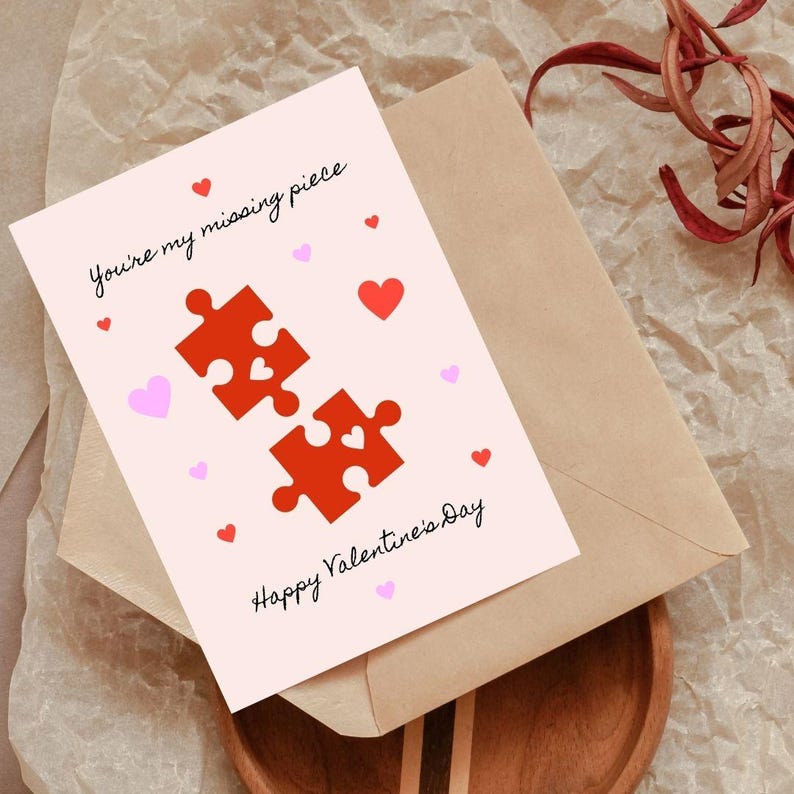 May include: A pink Valentine's Day card with the text "You're my missing piece" and "Happy Valentine's Day". The card features two red puzzle pieces that fit together, with a small heart in the center of each piece. There are small pink hearts scattered around the card.