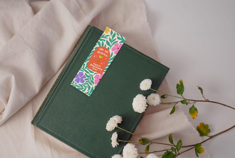 Puede incluir: Un libro verde con un marcap&aacute;ginas que dice "Eres hermosa. Recuerda siempre eso." El marcap&aacute;ginas tiene un dise&ntilde;o floral con flores rosas, amarillas y moradas.