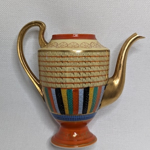 May include: A gold and orange ceramic teapot with a geometric pattern. The teapot has a wide base and a spout on the side. The pattern includes gold, orange, blue, green, and black colors.
