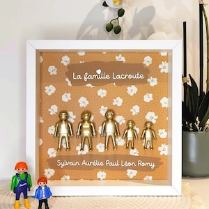 May include: A white square shadow box frame containing five gold-painted plastic figures arranged in a row against a mustard yellow background with a white floral pattern. The text "La famille Lacroute" and "Sylvain Aur&eacute;lie Paul L&eacute;on Romy" is printed on the background.