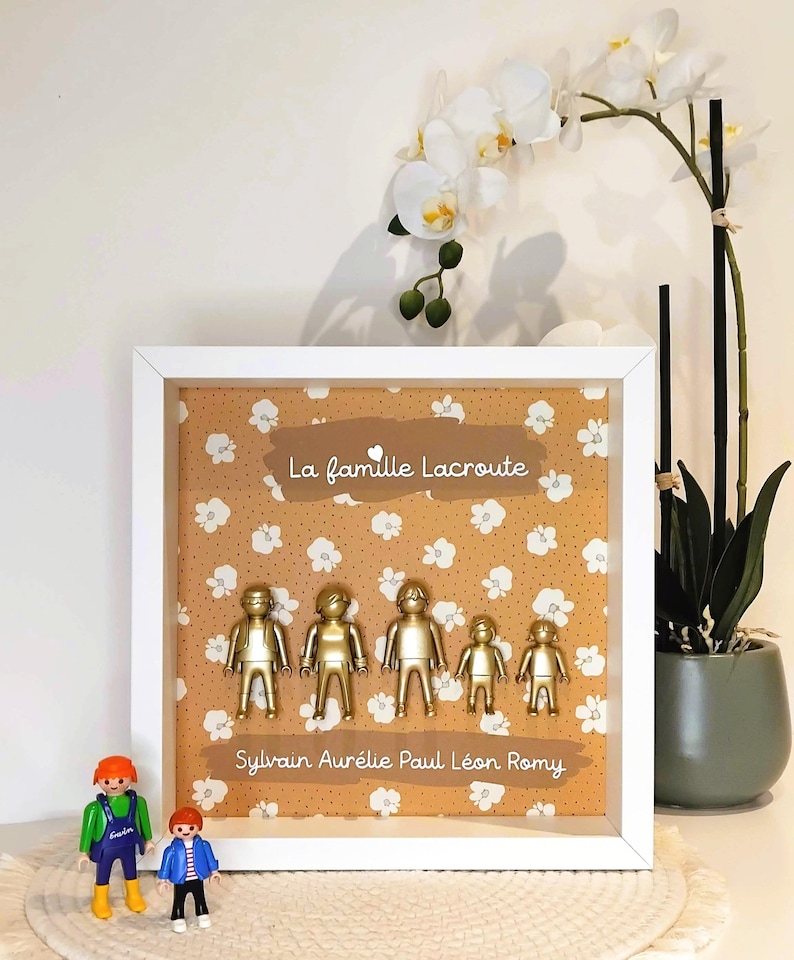 May include: A white square shadow box frame containing five gold-painted plastic figures arranged in a row against a mustard yellow background with a white floral pattern. The text "La famille Lacroute" and "Sylvain Aur&eacute;lie Paul L&eacute;on Romy" is printed on the background.
