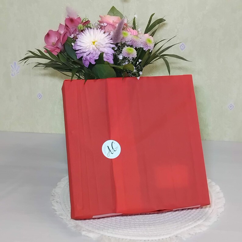 May include: A square package wrapped in vibrant red tissue paper, secured with a circular white sticker featuring the black letters AC. Behind the package sits a bouquet of flowers including pink roses, a large white chrysanthemum, and green foliage.
