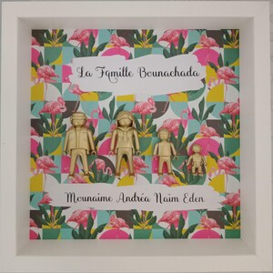 May include: A white shadow box frame containing four gold-colored plastic figures of varying sizes arranged in a row. The background features a repeating pattern of pink flamingos and green leaves. Text reads La Famille Bounachada and Mounaime Andr&eacute;a Na&iuml;m Eden.