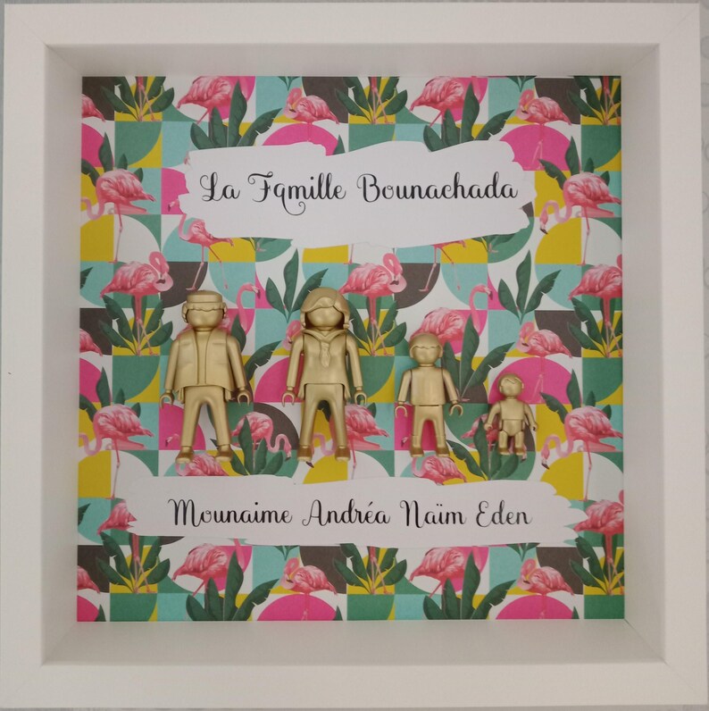 May include: A white shadow box frame containing four gold-colored plastic figures of varying sizes arranged in a row. The background features a repeating pattern of pink flamingos and green leaves. Text reads La Famille Bounachada and Mounaime Andr&eacute;a Na&iuml;m Eden.