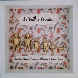 May include: A white square shadow box frame containing six gold-colored plastic figures and a dog figurine. The background features a pattern of pink and gray shapes with black floral line art. Text reads La Famille Aubrion and Antoine Ana&iuml;s Cl&eacute;mence Martin Simon Cyrus.