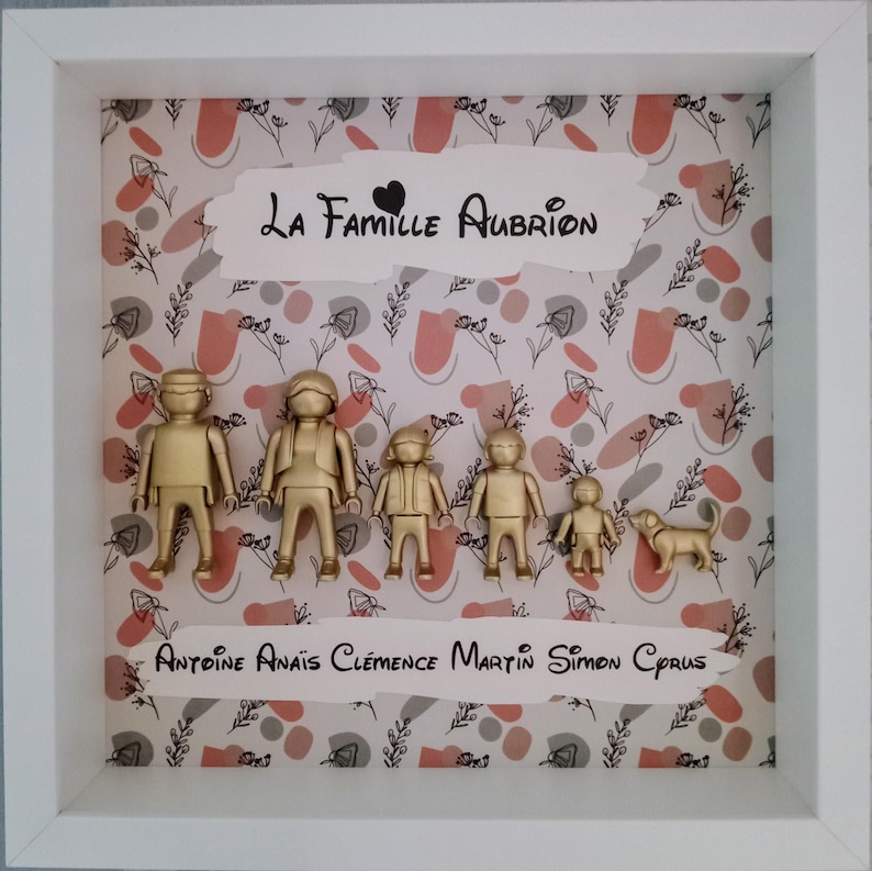 May include: A white square shadow box frame containing six gold-colored plastic figures and a dog figurine. The background features a pattern of pink and gray shapes with black floral line art. Text reads La Famille Aubrion and Antoine Ana&iuml;s Cl&eacute;mence Martin Simon Cyrus.