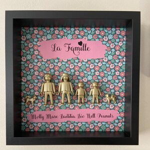 May include: A black shadow box frame containing a floral background with pink and teal flowers. Centered are four gold-colored plastic figures and two gold-colored dog figures. The text "La Famille" and "Molly Marc La&euml;titia L&eacute;o Nell Peanuts" appear on pink banners.