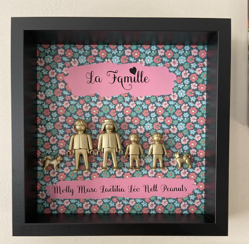 May include: A black shadow box frame containing a floral background with pink and teal flowers. Centered are four gold-colored plastic figures and two gold-colored dog figures. The text "La Famille" and "Molly Marc La&euml;titia L&eacute;o Nell Peanuts" appear on pink banners.